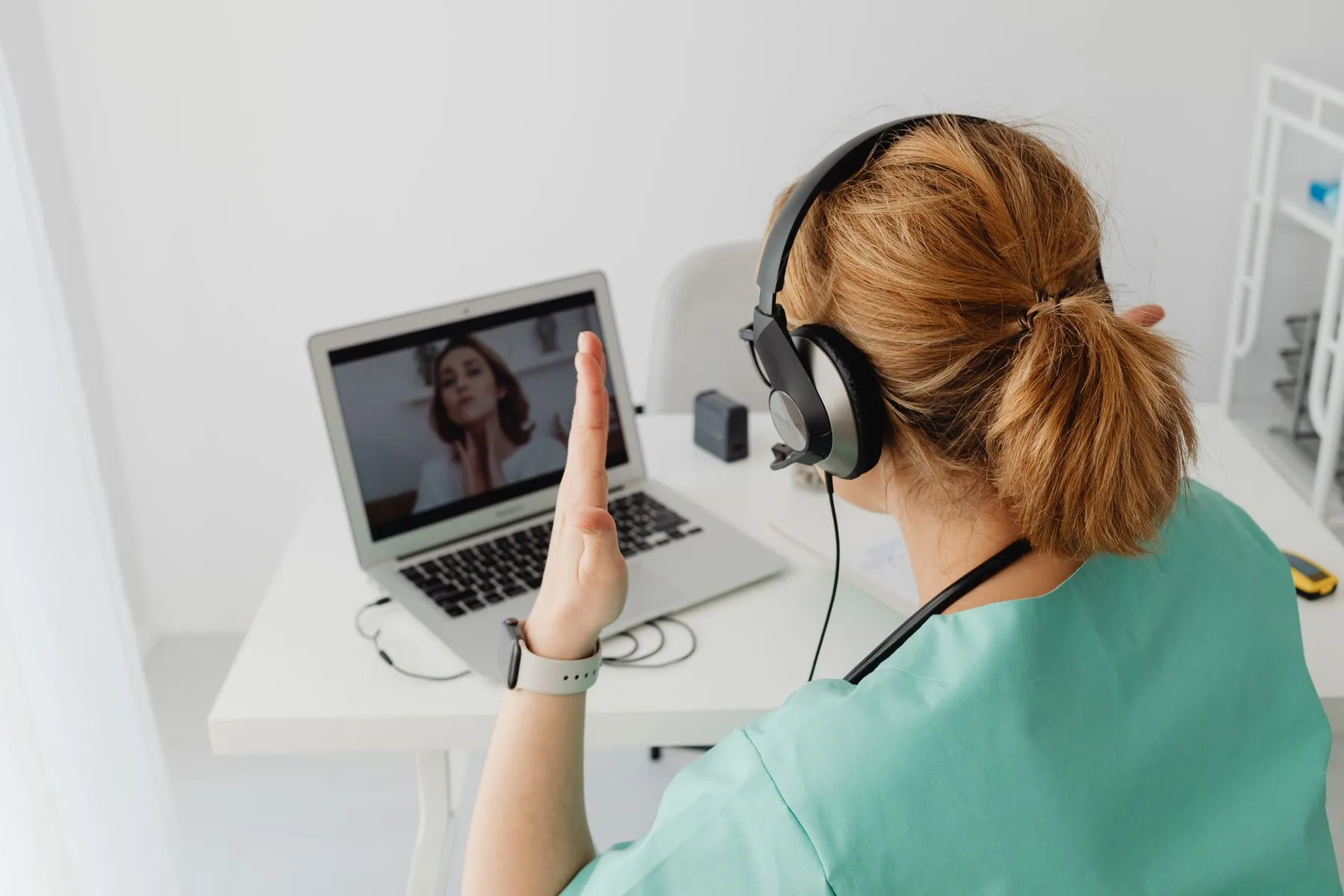 A clinician in teal scrubs and a headset waves to a patient on a laptop video call in a bright consultation room.